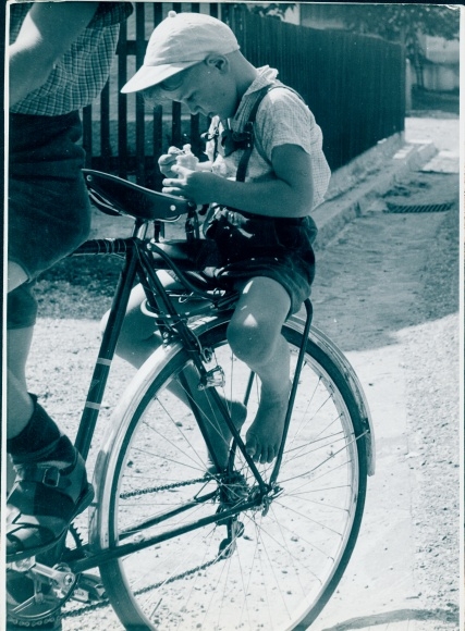 Photo of a child on a bicycle rack, wearing leather trousers and a peaked cap